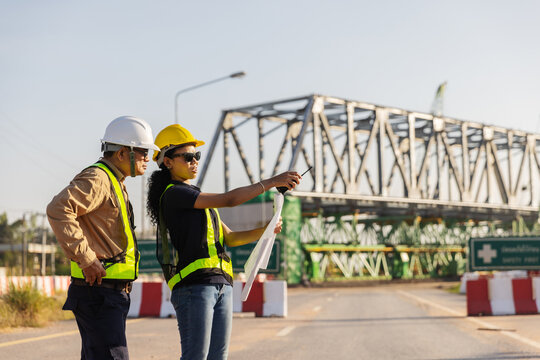 Two engineers in hard hats and reflective safety vests review construction plans, with one pointing toward a steel railway overpass under construction above a road.