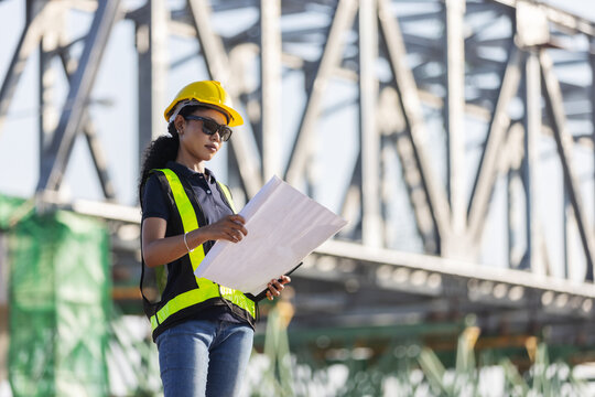 An engineer wearing a yellow hard hat and reflective safety vest reviews construction plans at a railway bridge site, with a steel truss structure in the background.