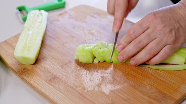 Woman hands hold a peeled cucumber steady and slice it lengthwise then crosswise into small pieces on a bamboo cutting board, a step by step hydration rich summer snack idea.