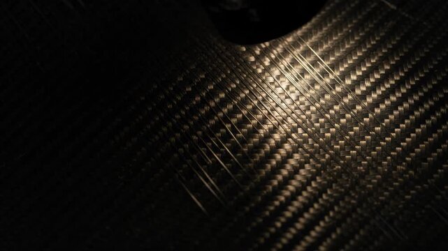 Dark abstract carbon fiber weave texture surface illuminated spotlight highlight pattern material background evoking mystery intrigue golden wallpaper line contrast warm light sleek aesthetic backdrop