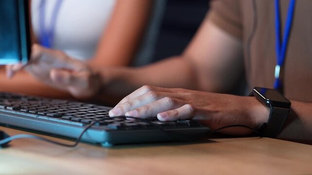 Close-up of hands typing on a computer keyboard at a wooden desk in an office, with a smartwatch on the wrist and a colleague working beside in the background.