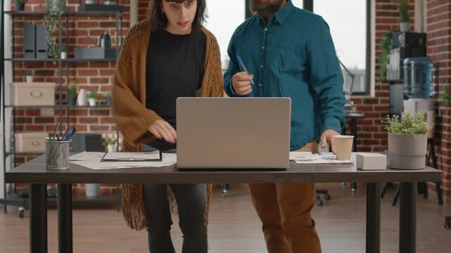 Laptop, collaboration and review as a woman and male colleague lean toward the same screen at an office table. Printed charts support joint data analysis, digital workflow and project planning.