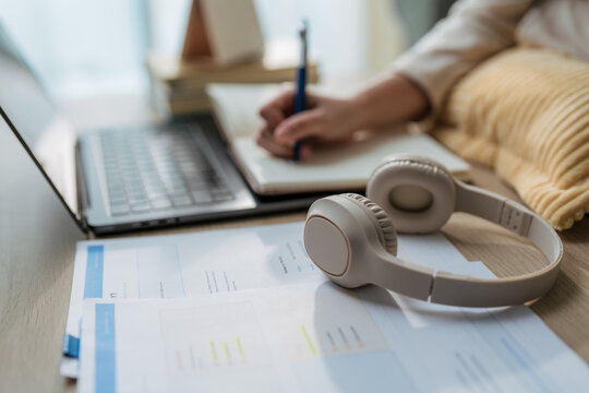 Close up of wireless headphones on desk with documents while blurred woman writing in notebook and using laptop in background for online learning, webinar, or remote working concept.