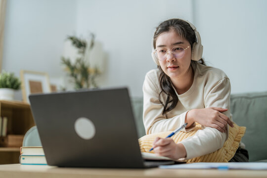 Focused young Asian woman wearing headphones using laptop and writing in notebook at home, representing online education, e-learning platform, webinar, and remote working lifestyle.