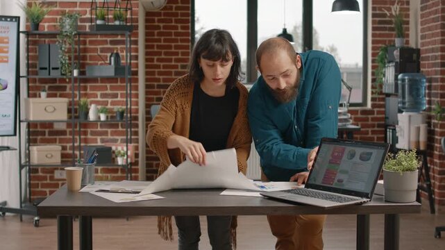 Charts, research and collaboration as two colleagues lean over printed diagrams while a laptop displays analytics. They discuss project results, compare figures and plan business next steps.