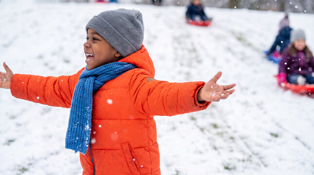 African child jumping to catch snowflakes in orange coat and blue scarf, laughing on a snowy hill with sled tracks for energetic winter posters.
