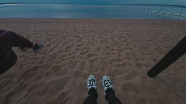 POV of people swinging above a sandy lakeshore, with legs and sneakers visible in motion as the swing moves toward calm blue water beneath a cloudy sky showing a moment of carefree adulthood.
