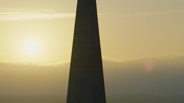 Aerial view moving upward along the Salisbury Cathedral spire to reveal its cross against a golden hazy sky with the sun glowing to the left.