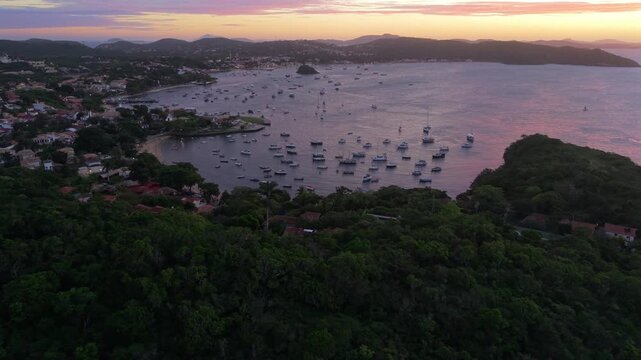 Drone flies forward and descends over vegetated hillside neighborhood towards calm bay filled with moored sailboats at Praia dos Ossos in Buzios under pink sunset skies.