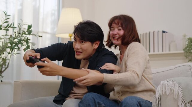 Happy Asian couple laugh together on a sofa as a woman playfully covers her partner's eyes while he holds a video game controller, capturing joyful gaming, affection and shared leisure at home.