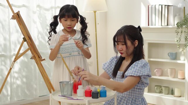 Asian mother and young daughter prepare colorful paints and brushes beside an easel, enjoying a hands-on art activity that encourages creativity, learning and family participation.