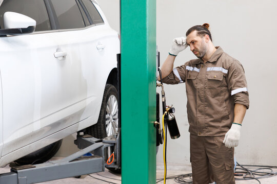 A tired mechanic feeling headache while leaning against a car lift in a garage.