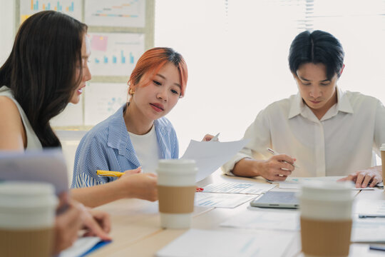 Young Asian colleagues reviewing documents and taking notes at conference table with coffee cups while discussing business reports in bright office meeting