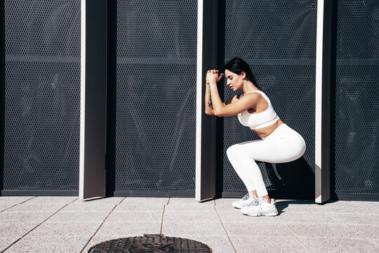 Fitness woman in sports clothing. Sexy young beautiful model athlete doing fitness workout. Female making exercises in street. Stretching out before training. Posing near urban wall. Makes squats
