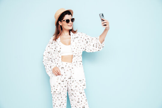 Young beautiful smiling female in trendy summer costume clothes. Carefree woman posing near blue wall in studio. Positive model having fun indoors. Cheerful and happy. In hat. Takes selfie photos