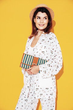 Young beautiful smiling female in trendy summer costume clothes. Carefree woman posing near yellow wall in studio. Positive model having fun indoors. Cheerful and happy. In hat. Holds clutch