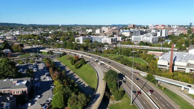 Curving interstate ramps and traffic sweep past a brick smokestack, parking lots and offices toward the University of Tennessee campus and hills in Knoxville, Tennessee. Aerial view