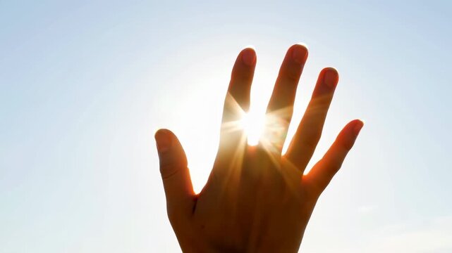 Open hand shifts gently with fingers spread against clear blue sky, allowing bright sunlight to shine through gaps and cast warm glowing edges around fingers in outdoor closeup