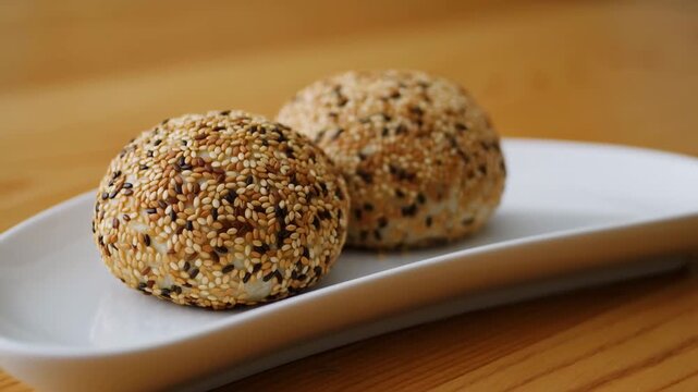 Seed-coated bread rolls rest on white serving platter on wooden table as close view gradually tightens, highlighting golden crust and contrasting pale and dark seeds in warm light