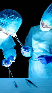Medical professionals performing surgery in sterile operating room, hands wearing blue gloves, holding instruments material, demonstrating precision focus action, dark background, vertical photo.