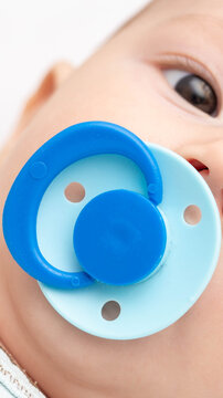 Close-up of a baby face with blue pacifier in mouth, partial view of eye, peaceful expression, child comfort item, infant accessory, soft skin detail, young human, vertical photo