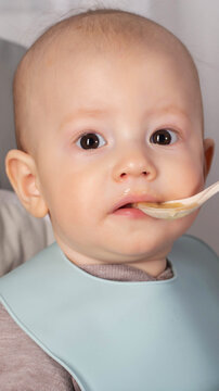 Close-up portrait of young infant with wide eyes being spoon-fed nutritious puree, wearing blue bib, mouth open slightly for food intake, healthy development focus vertical photo
