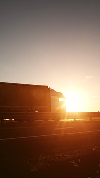 Heavy semi-trailer truck travels on open highway road at golden hour sunset, sun flare illuminates vehicle and road barrier, symbolizing long distance freight transport journey, vertical photo
