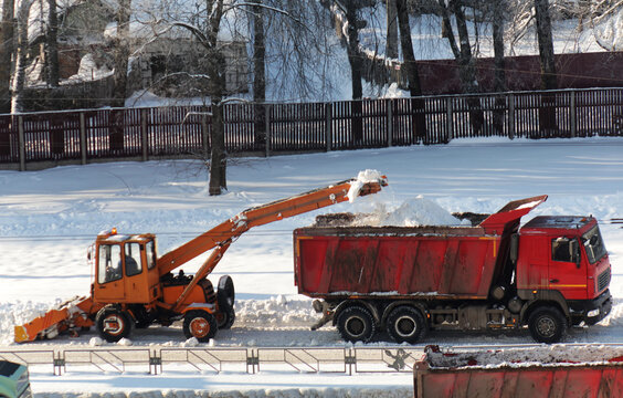 Orange snow loader machine transfers fresh white snow into red dump truck for transport during sunny winter day outdoors.