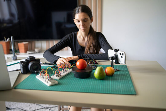 Female engineer using tablet to program smartphone AI camera recognition for sorting fruits on a tech lab desk.