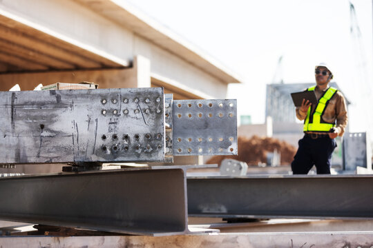 Close-up of structural steel beams and drilled connection plates prepared for bridge construction, with an engineer holding a tablet softly blurred in the background.