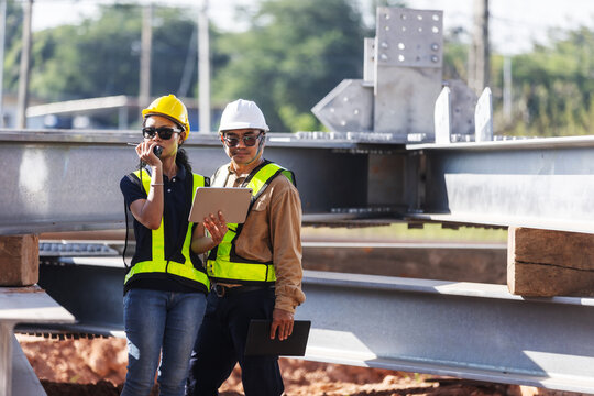 Two engineers wearing hard hats and reflective safety vests inspect structural steel beams at an outdoor construction site, using a tablet and handheld radio to coordinate quality checks.