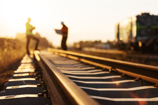 Low angle view of steel railway rails, concrete sleepers and fastening clips in warm evening light, with engineers and railway machinery softly blurred in the background.