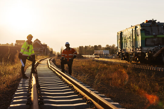 Two engineers in reflective workwear coordinate railway construction in warm evening light. One uses a handheld radio while the other reviews plans.