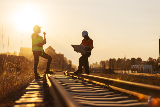 Two engineers in reflective workwear coordinate railway construction in warm evening light. One uses a handheld radio while the other reviews plans