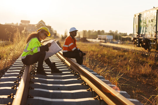 An engineer in reflective workwear and a hard hat reviews documents while seated on a railway rail, with a colleague nearby and rail equipment in the background under warm evening light.