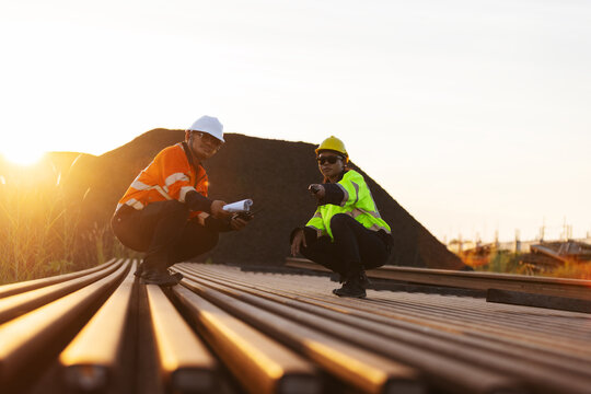 Two engineers in hard hats and reflective workwear inspect steel railway rails at an outdoor site. One checks the rail surface with a handheld tool while the other records findings on a clipboard.