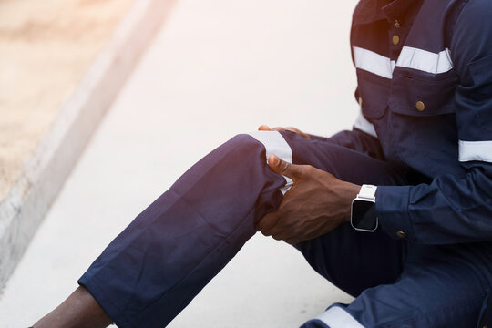 African American industrial worker sitting outdoor holding injured knee in severe pain after workplace accident on construction site