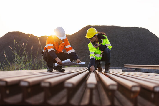 Two engineers in hard hats and reflective workwear inspect steel railway rails at an outdoor site. One checks the rail surface with a handheld tool while the other records findings on a clipboard.