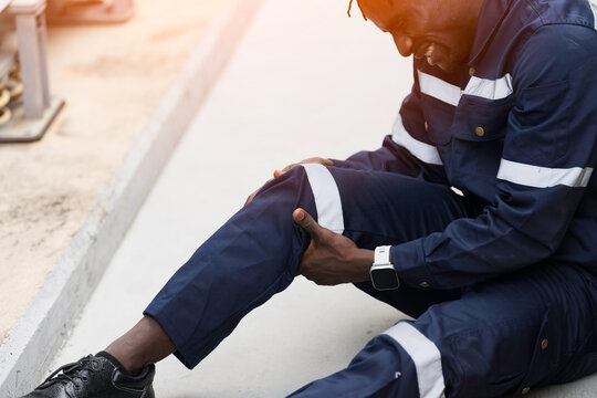 African American industrial worker sitting outdoor holding injured knee in severe pain after workplace accident on construction site