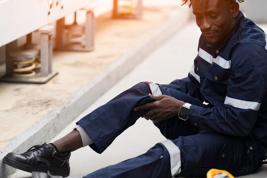 African American industrial worker sitting outdoor holding injured knee in severe pain after workplace accident on construction site