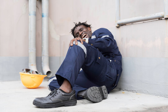 African American industrial worker sitting outdoor holding injured knee in severe pain after workplace accident on construction site