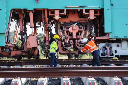 Two engineers wearing hard hats and reflective workwear inspect exposed mechanical components beneath a railway track machine, with steel rails and concrete sleepers in the foreground.