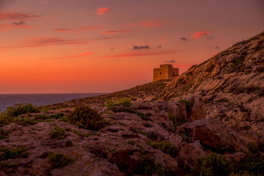 Dwejra Coastal Slopes and Ancient Stone Watchtower Silhouetted Against a Vibrant Orange and Pink Sunset Sky, Gozo Island, Malta