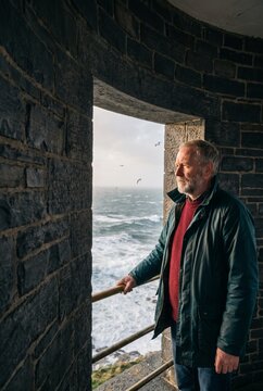 Thoughtful mature bearded man standing inside a historic stone tower looking out over a rough ocean with crashing waves and flying seagulls