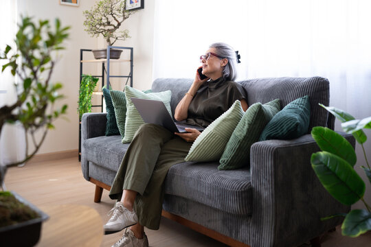 Senior white woman calling on phone and using laptop on gray sofa