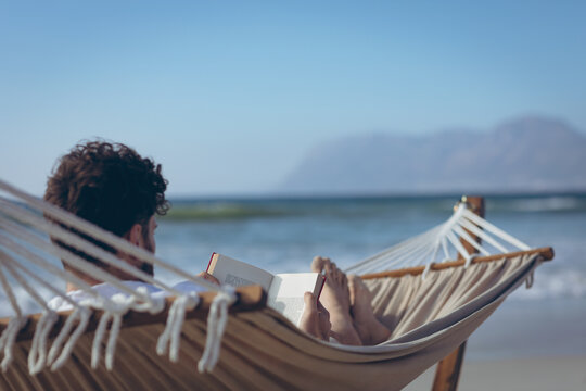 Man reading book while lying on hammock at beach