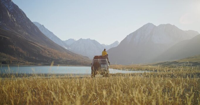 Man in a yellow jacket speaking on the phone while sitting on the roof of a parked car near a calm mountain lake with snowy peaks in the background. Warm light, golden meadow, and open alpine