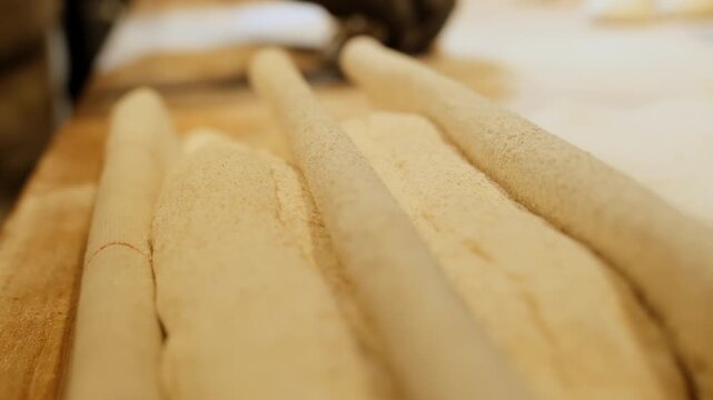 Baker in black gloves shapes long dough ropes on floured wooden table. Skilled worker aligns soft pastry strips forming even pieces
