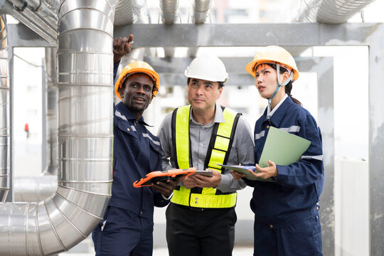 Group of engineer and technician discussing maintenance check, inspecting and reviewing project plans and safety checks beside metal piping system on building roof at construction site