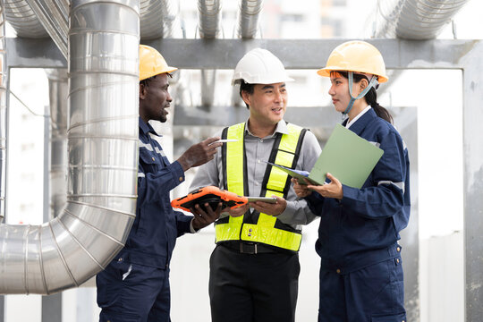 Group of engineer and technician discussing maintenance check, inspecting and reviewing project plans and safety checks beside metal piping system on building roof at construction site